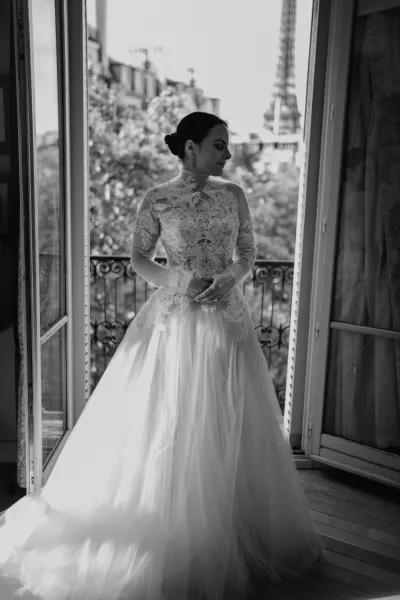 Bridal portrait in black and white of a bride by window light, wearing a long sleeve lace dress, looking away beside a balcony railing