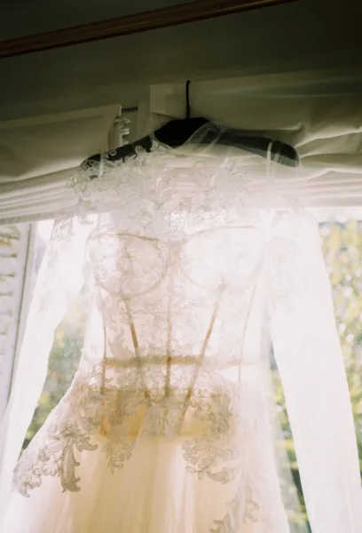 Wedding dress with lace appliqué corset bodice and sheer sleeves on a hanger, softly lit by window light with greenery outside