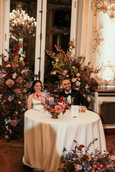 Sweetheart table with wedding sweetheart table decor, lush florals, candles and wine glasses beneath a chandelier in an ornate ballroom