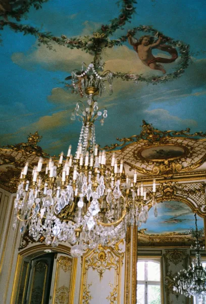 Ornate chandelier with crystal accents and candle-style bulbs, hanging beneath a cherub ceiling mural in a gilded ballroom interior