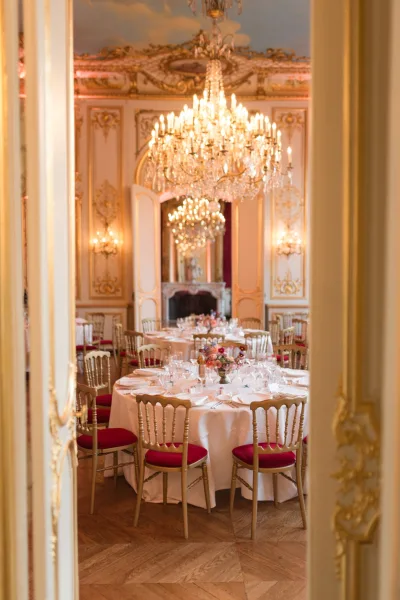 Reception tablescape with white tablecloths and floral centerpieces on round tables beneath a crystal chandelier in an ornate gilded ballroom