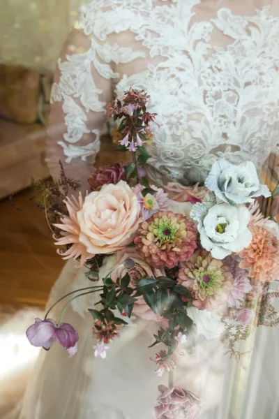 Bridal bouquet in garden style with blush rose, zinnia, dahlia, lisianthus and greenery, held against a lace wedding dress in soft indoor light