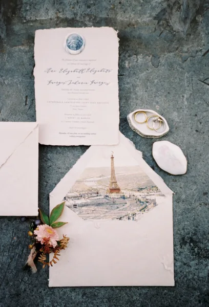 Invitation suite flat lay with deckle edge wedding invitation, envelope, rings, and blue wax seal on a gray stone surface