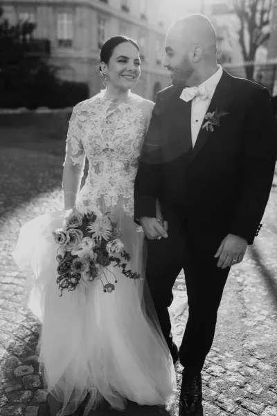 Couple portrait in black and white of bride in long-sleeve lace dress and veil walking arm in arm with groom in tuxedo beside a stone building