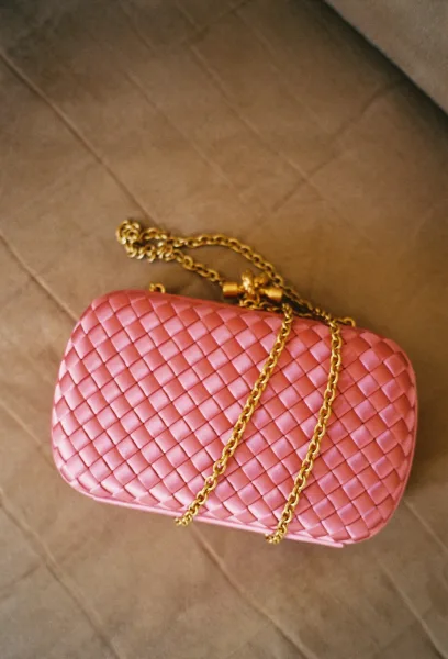Wedding clutch with pink woven texture and gold chain strap, featuring clasp closure, resting on a tan fabric surface
