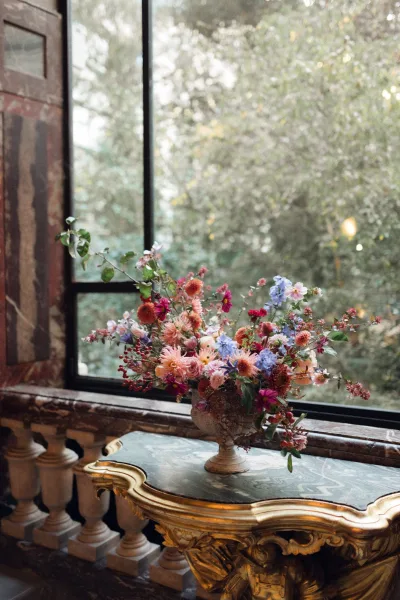 Wedding floral arrangement in a compote vase with colorful blooms, greenery and berry stems on a vintage console by a large window
