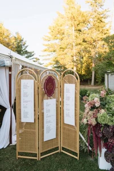 Wedding seating chart on a cane room divider with paper escort cards and hydrangea florals, set on a lawn under a clear tent with string lights