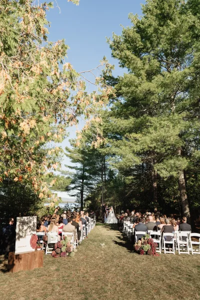 Outdoor wedding ceremony with white folding chairs lining a flowered aisle toward bride and groom by a lake, framed by pine trees under blue sky