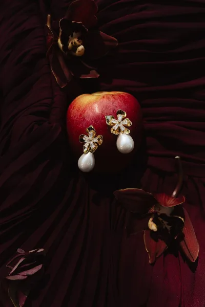 Wedding earrings with pearl drop earrings and crystal-studded gold floral design, styled with orchid and apple on burgundy fabric backdrop