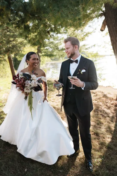 Couple portrait of bride and groom laughing, holding champagne coupes as she cradles a bouquet by a sunlit lakeside with pines