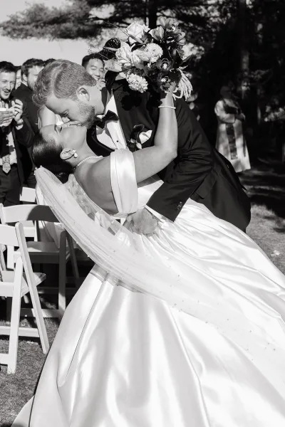 Wedding kiss portrait of groom dipping bride in off-the-shoulder gown and veil, bouquet raised, guests on lawn aisle with trees behind