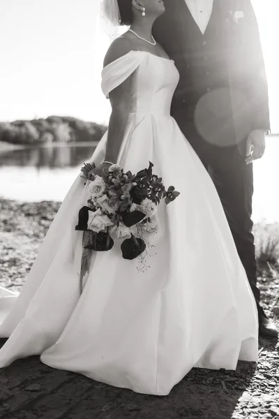 Couple portrait in a black and white wedding portrait, bride in veil holding bouquet beside groom in tuxedo by a lakeside walkway