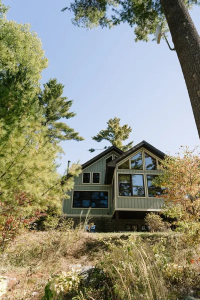 Woodland cabin exterior with large windows and gabled roof set on a hillside among pine trees and shrubs under a clear blue sky