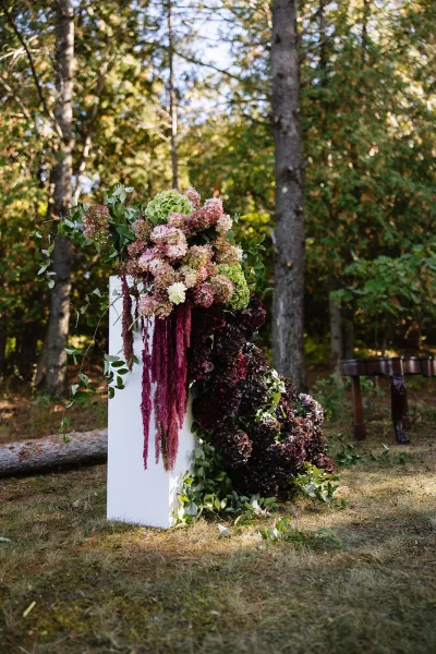 Ceremony floral arrangement on a wedding floral pedestal with hydrangeas and hanging amaranthus, set on a lawn among woodland trees