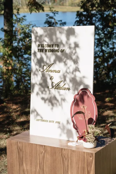 Wedding welcome sign with gold lettering on an acrylic panel atop a wood pedestal, set by a lake with trees in dappled sunlight
