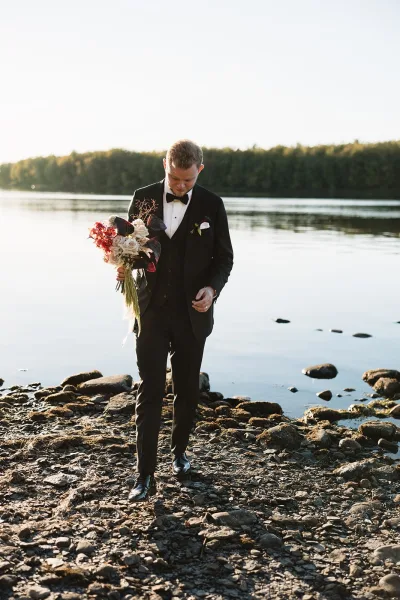 Groom portrait of a black tie groom holding bouquet, looking down in a black tuxedo with boutonniere beside a rocky lakeshore
