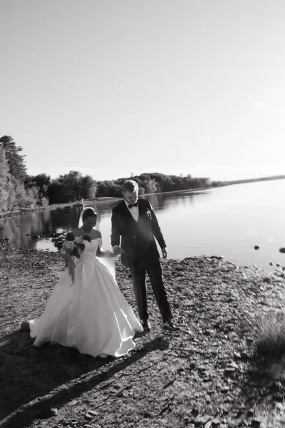 Couple portrait of bride and groom holding hands on a rocky lakeshore, her veil and bouquet trailing as he leads in tuxedo