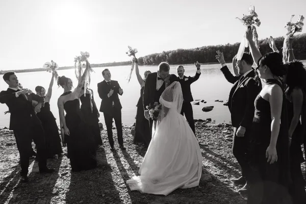 Wedding kiss moment as bride and groom kissing while wedding party cheers behind them by a sunset lakeshore with veil and bouquets