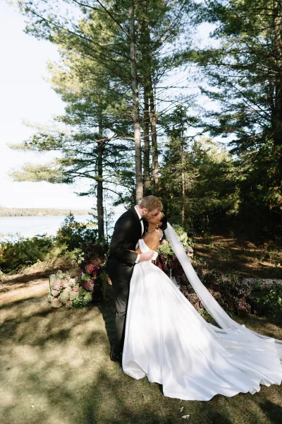 Wedding kiss portrait of bride and groom kissing in a dip, long veil flowing as they stand on a lakeside lawn with pine trees behind