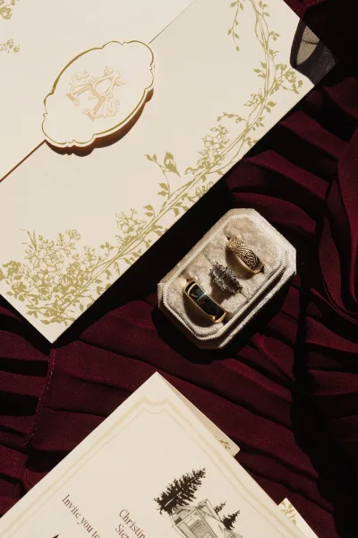 Wedding ring flatlay with a wedding invitation suite, velvet ring box, stacked bands, monogram seal, and ribbon on dark fabric backdrop