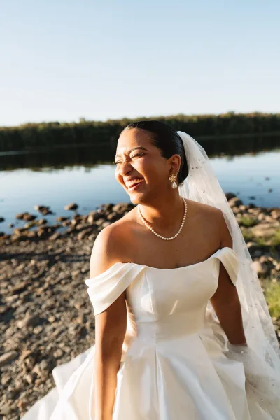 Bridal portrait of a laughing bride in an off the shoulder wedding dress with cathedral veil and pearls by a rocky lakeshore