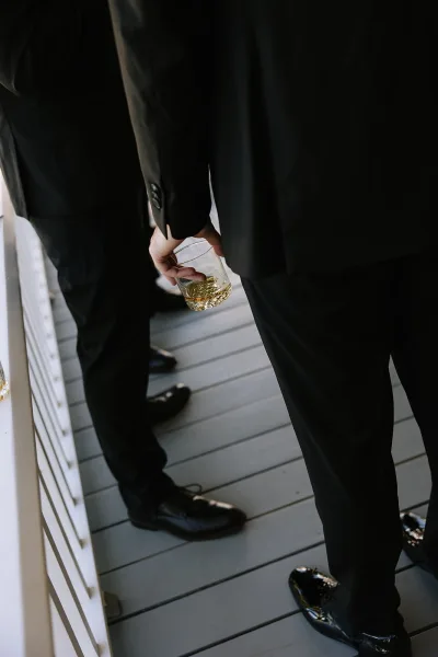 Groomsmen candid in black suits with a whiskey glass in hand, gathered by a porch railing on a wood deck floor pre-ceremony
