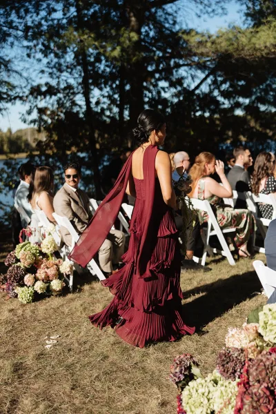 Wedding processional with bridesmaid walking down aisle in burgundy one-shoulder dress and chiffon scarf, holding bouquet at lakeside lawn ceremony