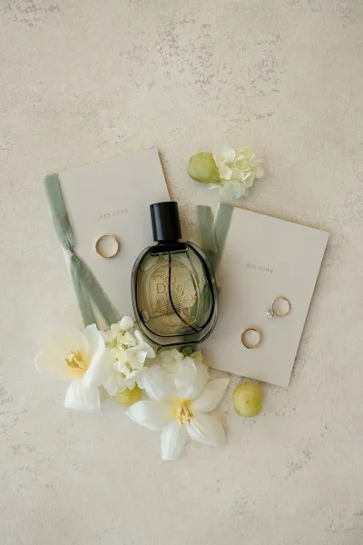Wedding flat lay featuring gold wedding bands with vow books, perfume bottle, silk ribbon, white flowers, and green grapes on a neutral textured surface