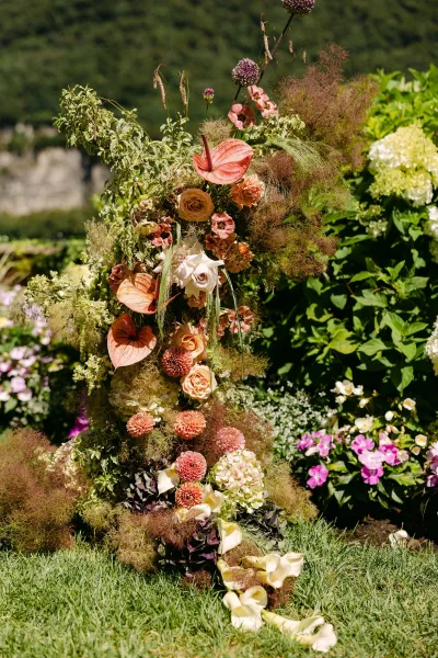 Ceremony floral arrangement of roses, anthuriums, dahlias, and hydrangea with wispy greenery on lawn beside garden shrubs and hillside greenery