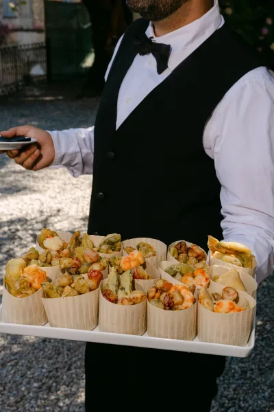 Wedding cocktail hour with passed appetizers as a server in a black vest and bow tie offers fried bites in paper cups on a tray outdoors