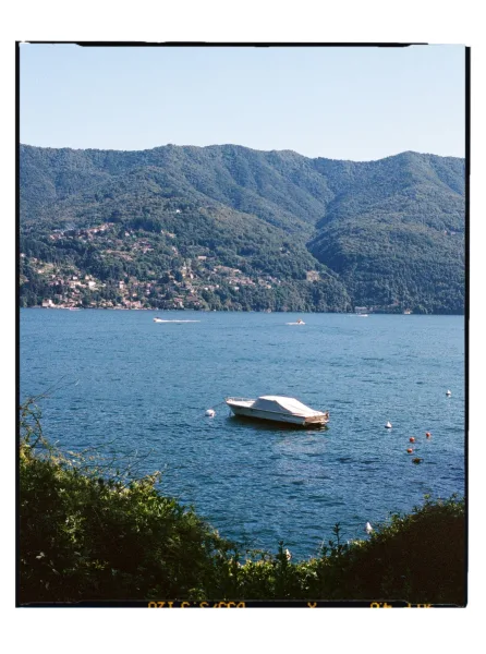 Lake boat view with a covered motorboat moored by buoy line on calm blue water, mountains and a hillside town in the distance