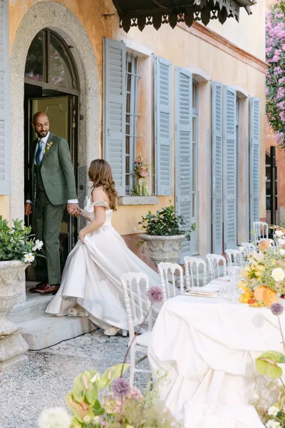 Wedding couple portrait of bride leading groom holding hands past a long white-linen banquet table in a villa courtyard with blue shutters