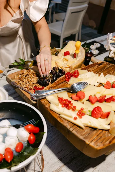 Wedding cheese board with fruit on a rustic wooden serving board, bridal hand with engagement ring reaching near grapes and cheeses on table