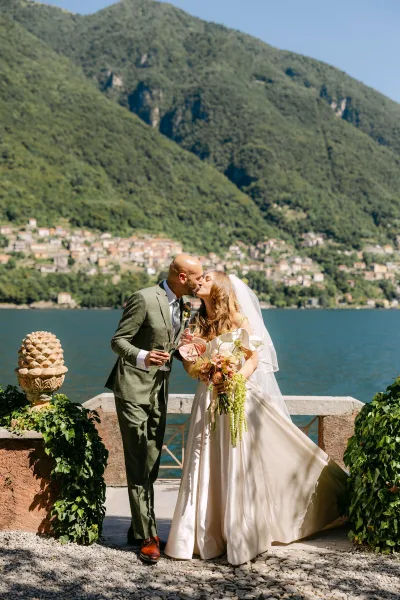 Wedding kiss as bride and groom toast with champagne flutes, her bouquet and veil on a stone terrace with lake and mountains behind