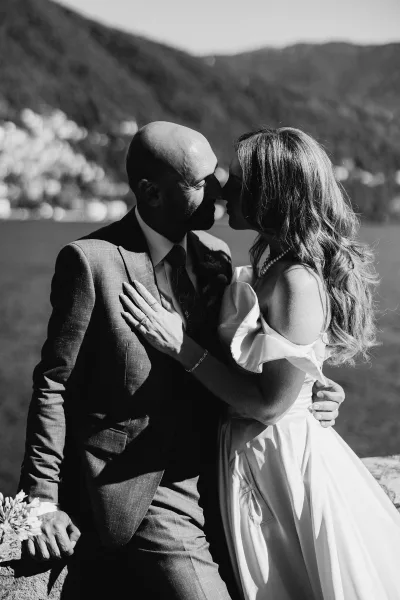 Wedding kiss portrait of bride and groom kissing as she grips his lapel showing her ring, with mountain lake backdrop on a stone ledge