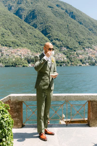Groom portrait with coffee cup in hand, wearing a green suit and sunglasses on a stone terrace overlooking a lake and mountains