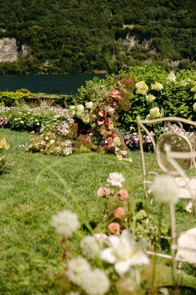Ceremony florals with a wedding ceremony floral installation of hydrangeas, roses, and greenery on lawn grass by a lake and hillside trees.