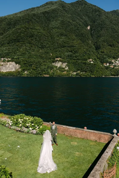 Wedding first look as bride walking to groom from behind, long veil and train flowing on lawn by stone wall with lake and mountains
