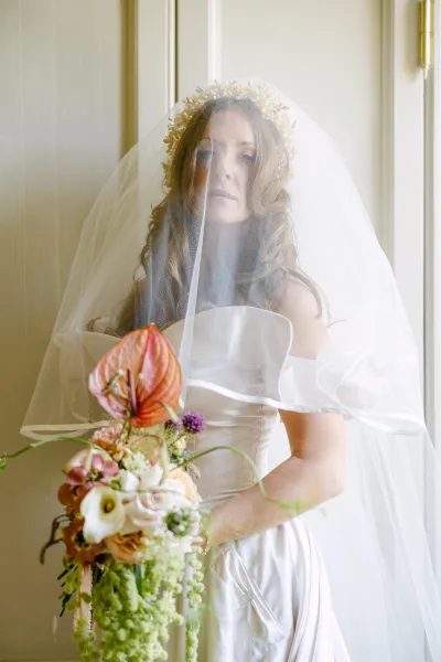 Bridal portrait with veil over face, wearing a strapless wedding dress and floral headpiece, holding anthurium and calla lily bouquet indoors