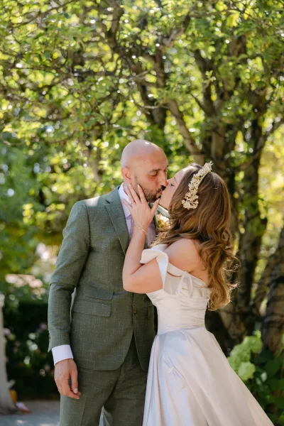 Wedding kiss as bride and groom kissing in sunlit garden trees, bride in off-shoulder dress touching his face with hair accessory and rings
