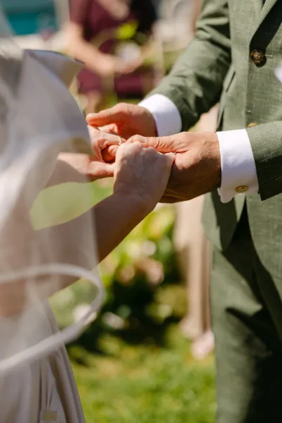 Ring exchange as groom places a wedding ring on the bride’s finger, veil edge visible, with garden greenery and blurred guests behind