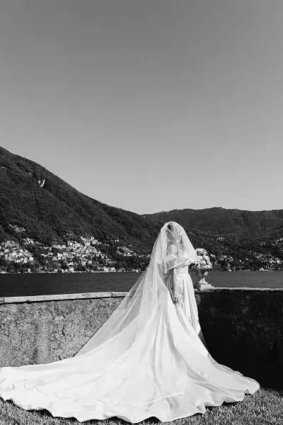 Bridal portrait in a black and white wedding portrait style, bride in strapless gown with long train and bouquet on a stone terrace by a lake and mountains.