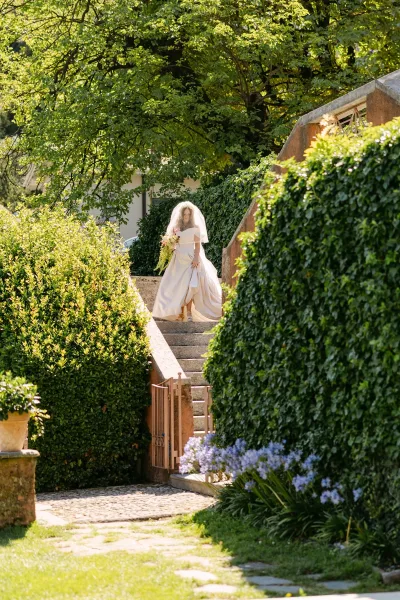 Bridal portrait of a bride walking down stone steps in a garden, wearing an off-the-shoulder gown and veil, holding a cascading bouquet