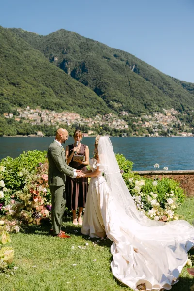 Wedding vows as bride and groom hold hands, her veil and long train flowing on a lakefront lawn with mountains behind