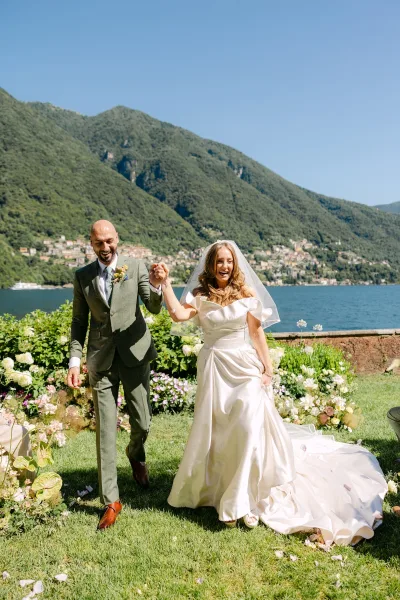 Recessional moment as newlyweds walk hand in hand, bride in off-shoulder gown and veil, groom in green suit by mountain lake backdrop