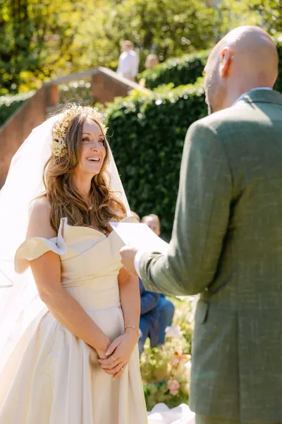 Wedding vows as groom reads from vow cards while bride smiles in a floral crown and veil during an outdoor wedding ceremony in sunny garden steps