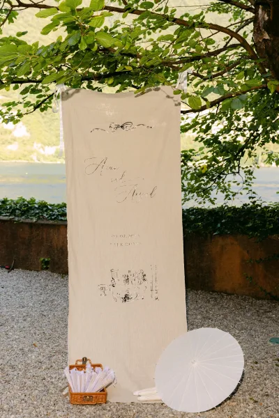 Wedding welcome sign with calligraphy wedding sign lettering on a fabric banner tied to a tree above a wicker basket of lace fans by a lake