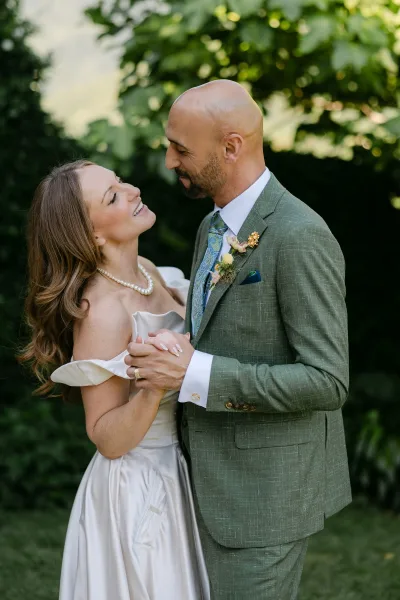 Couple portrait of bride and groom holding hands, she looks up at him in an off-shoulder dress with pearls amid garden greenery