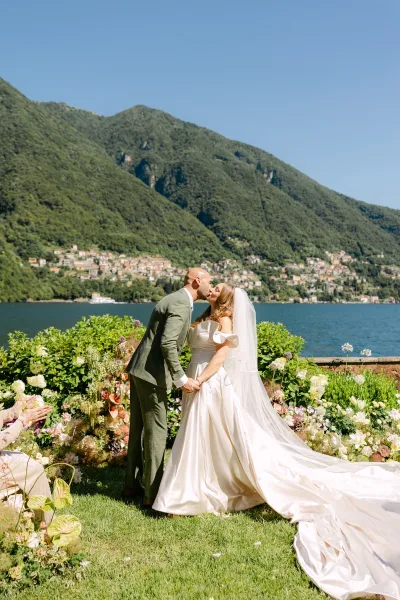 Wedding kiss portrait of bride and groom holding hands, her veil and long train flowing by aisle flowers with lake and mountains beyond