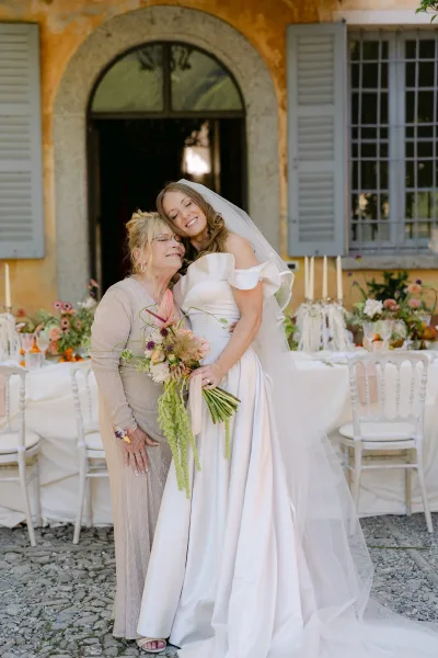 Bride with mother embraces in front of a stucco building with arched doorway, holding a ribbon-wrapped bouquet and wearing a veil